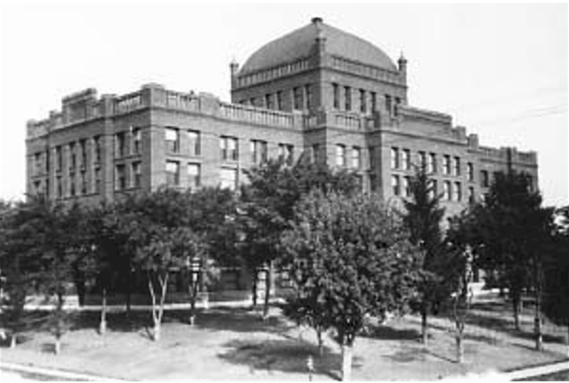 Black-and-white photograph of a large historic brick government building behind trees